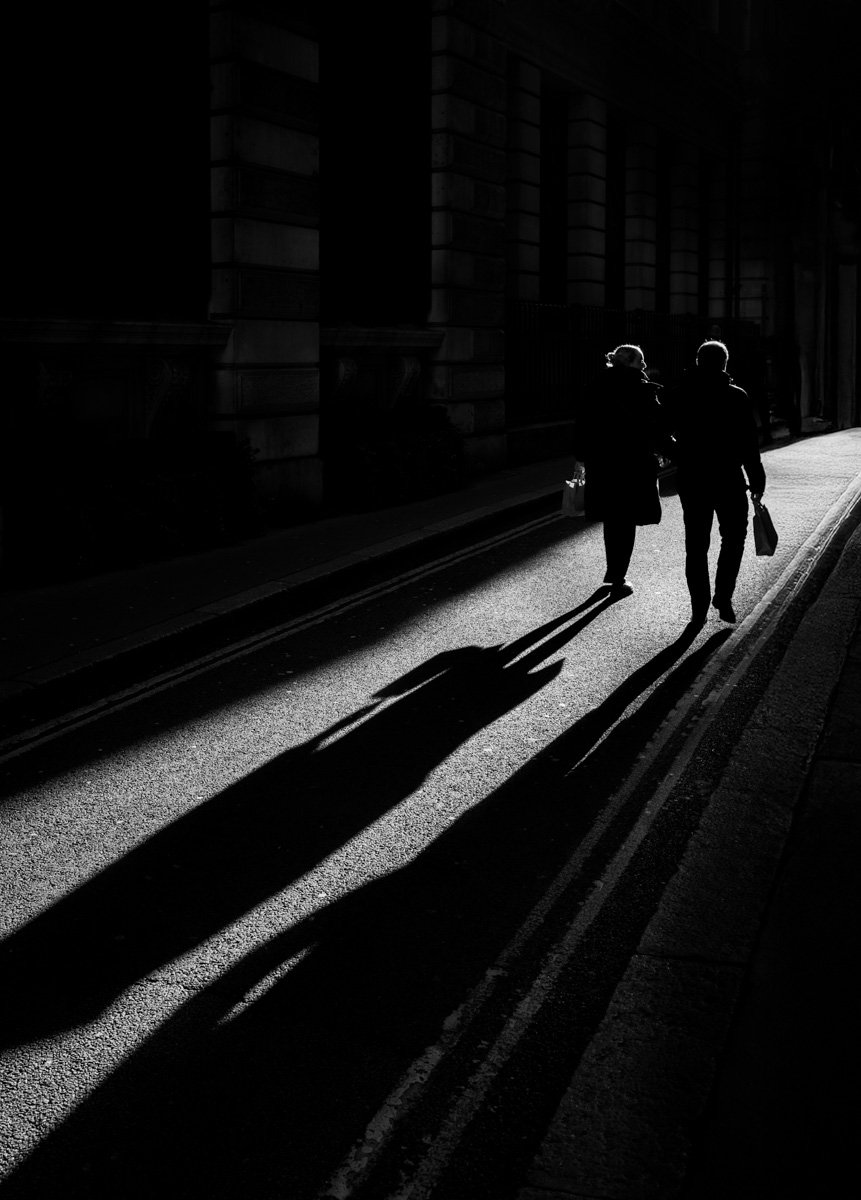 Two pedestrians walking through strong sunlight with long shadows on the street.