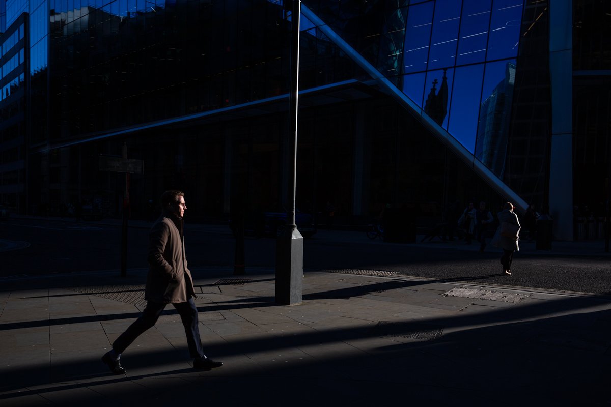 Man walking through a strip of sunlight beside a blue glass city building.