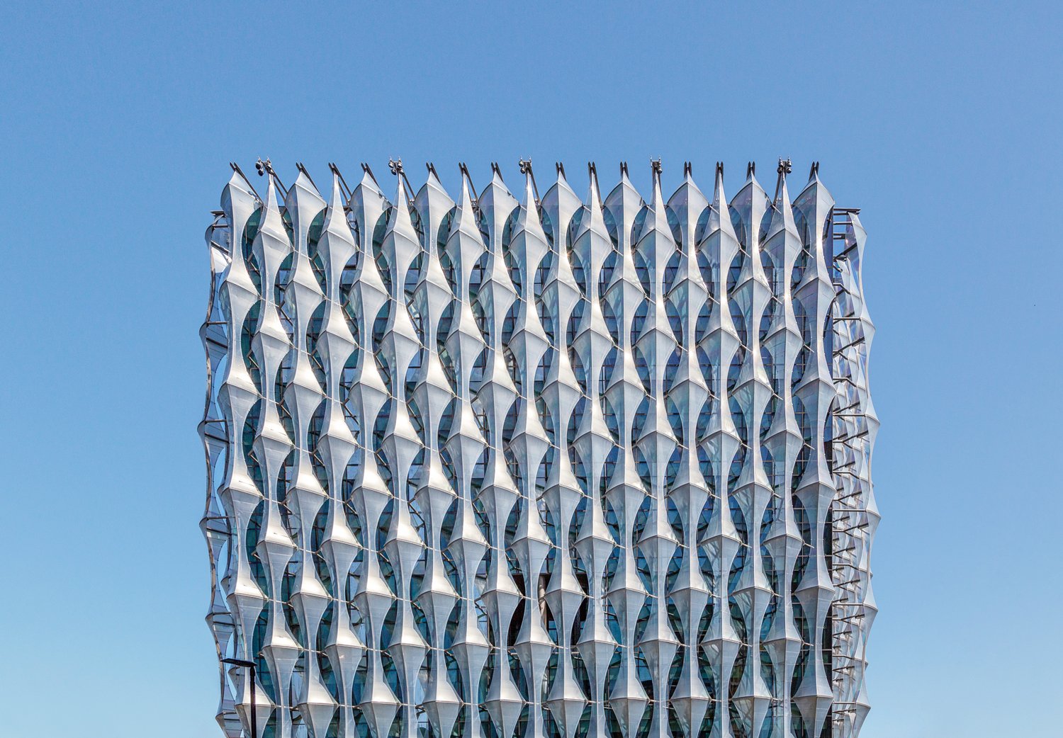 Repeating silver geometric facade on a tower against blue sky.