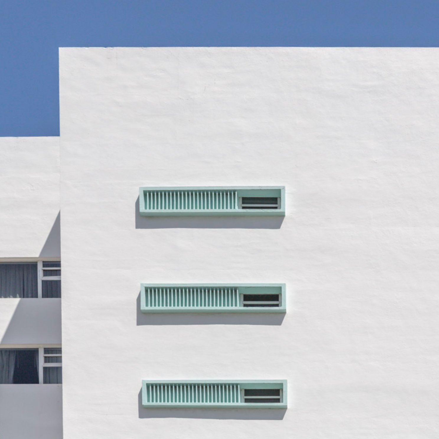 Minimal white facade with a small green louvred opening beneath blue sky.