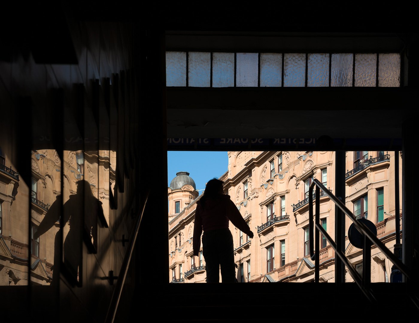 Silhouetted figure on steps framed by a doorway with sunlit city architecture beyond.