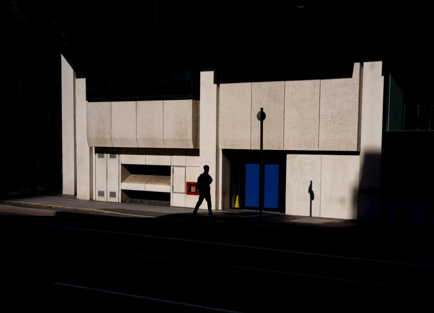 Lone pedestrian crossing a pool of sunlight in front of a pale modernist building.