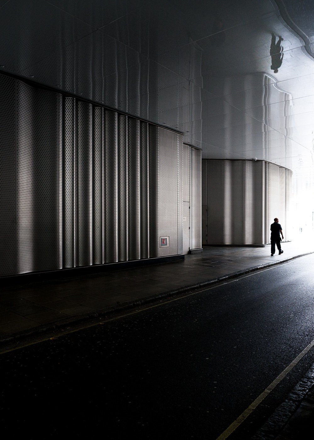 Lone pedestrian walking beside a corrugated metal facade at the edge of bright light.