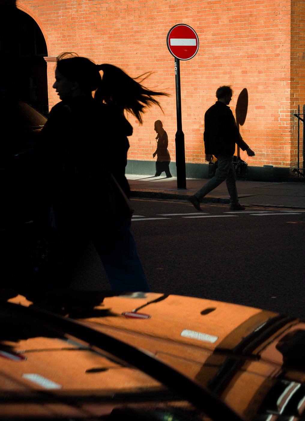 Layered pedestrians crossing bands of sunlight and shadow beside warm brickwork.