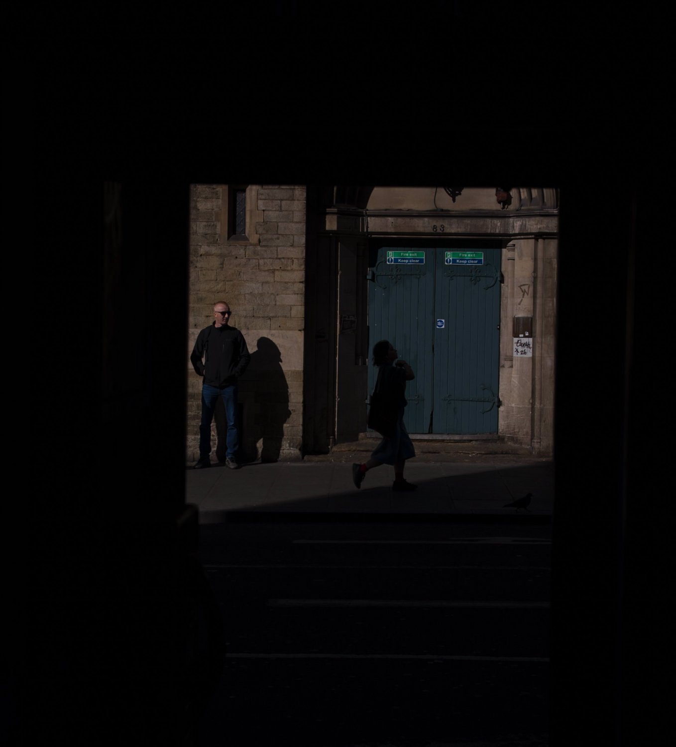 Man standing beside a stone wall while a woman crosses in front of blue doors.