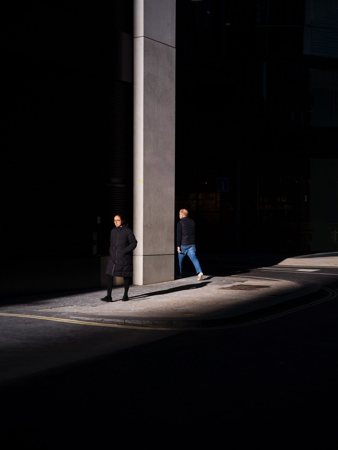 Two pedestrians separated by a tall concrete column in a narrow pool of sunlight.