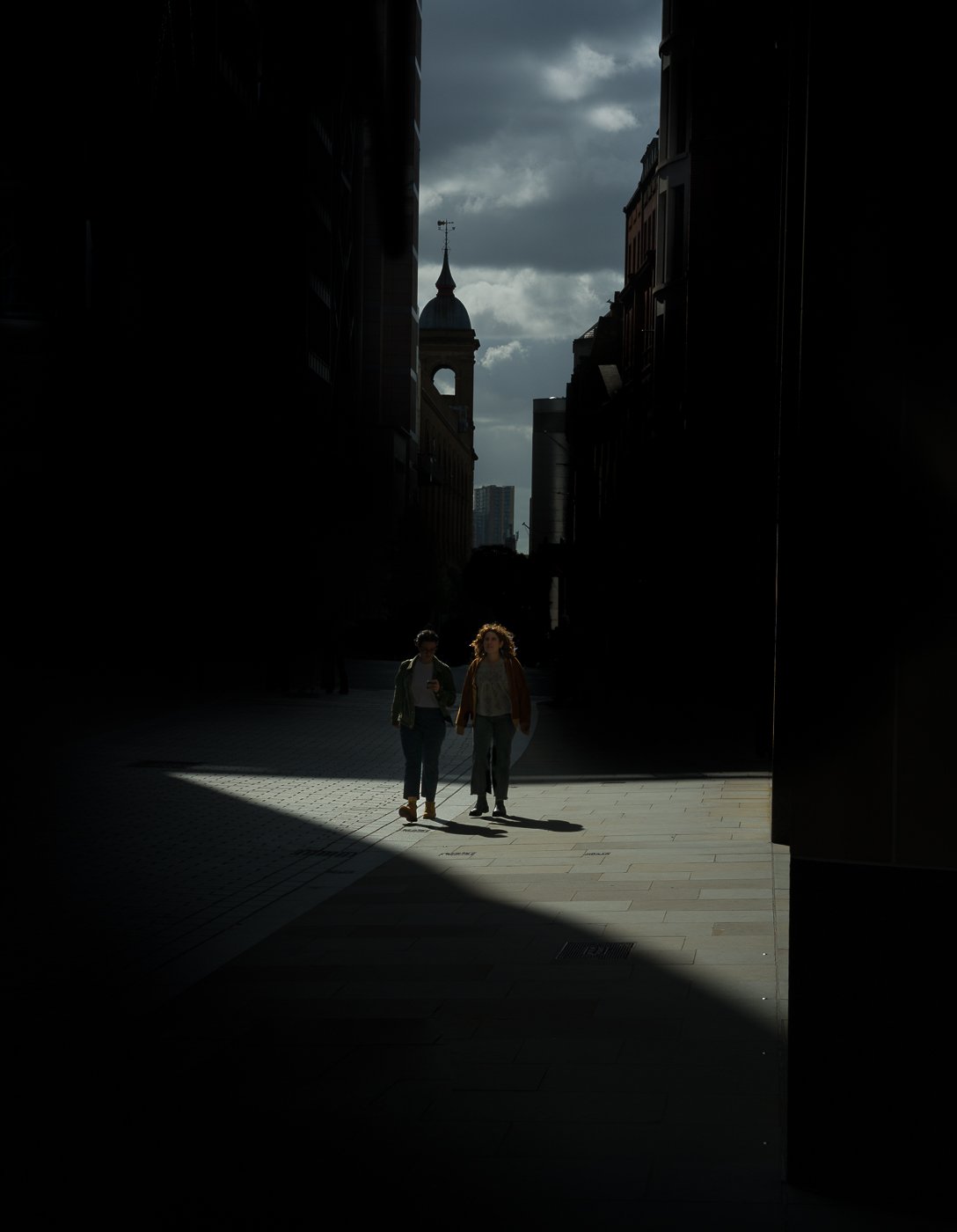 Two people standing in a patch of sunlight between dark city buildings.