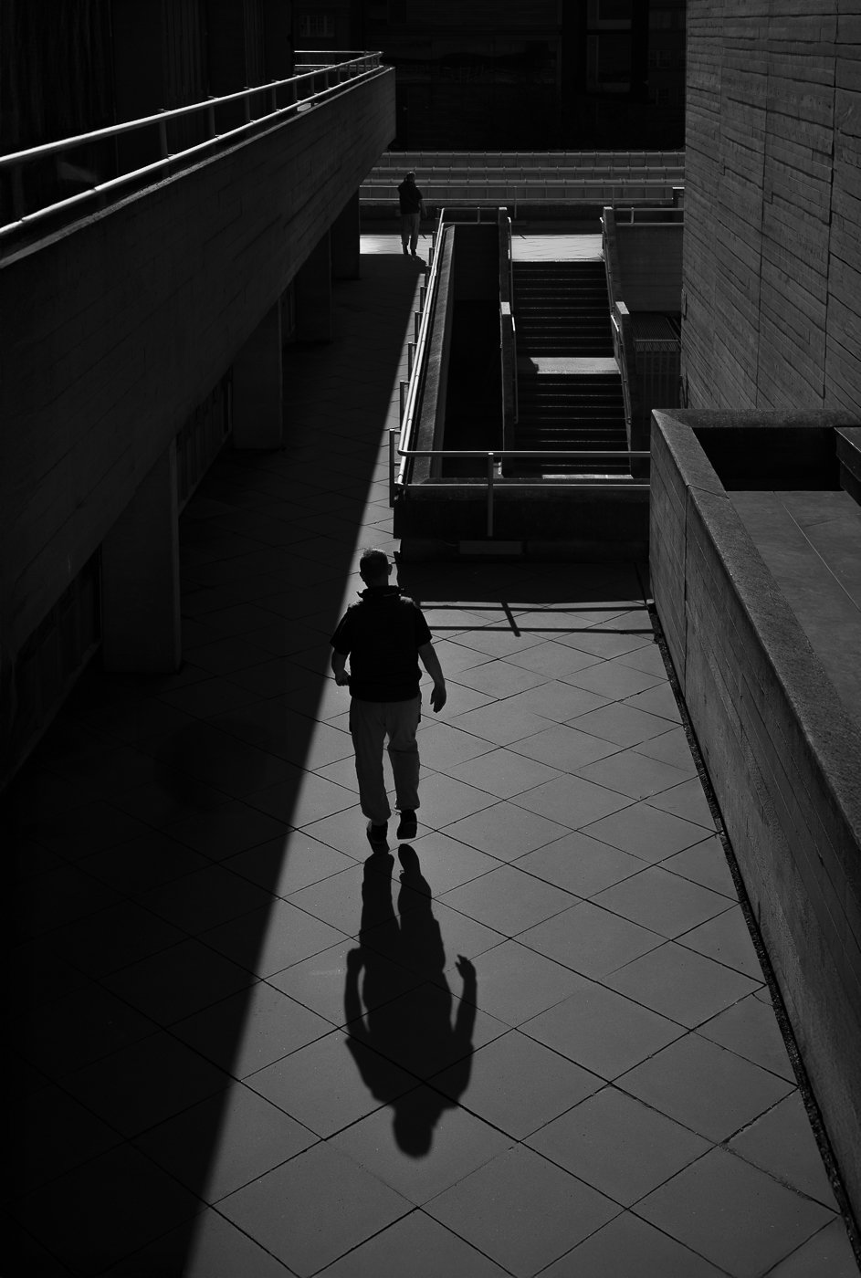 Pedestrian seen from above crossing geometric paving between stairs and deep shadow.